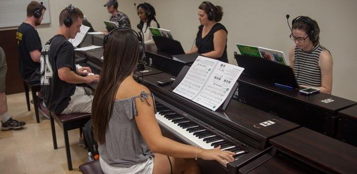 Eight pianos back to back down the middle of a classroom with a student playing and wearing headphones at each one.