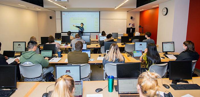 view from the back of the room of Dr. Haleh Khojasteh's Computer Science I class where she points at content on a projector screen in front of rows of students at desks with laptops open 